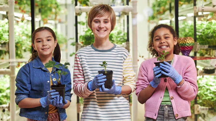 Three diverse kids, Caucasian boy, girl, Black girl, standing in greenhouse holding potted plants and smiling, wearing blue gloves, participating in gardening activity together.